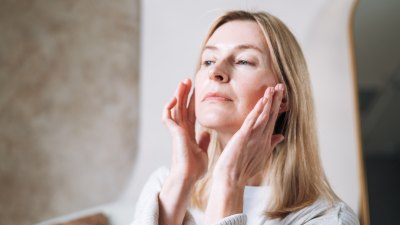 امرأة بالغة مركزة تمارس التدليك الذاتي للوجه في حمامها المضاء جيدًا. باستخدام أطراف أصابعها، تقوم بتنعيم بشرتها بلطف لتحسين الدورة الدموية وتقليل التوتر وتعزيز التوهج الصحي والطبيعي. تجسد لحظة العافية الأصيلة هذه في المنزل اتجاه الجمال والعناية الذاتية الشائع